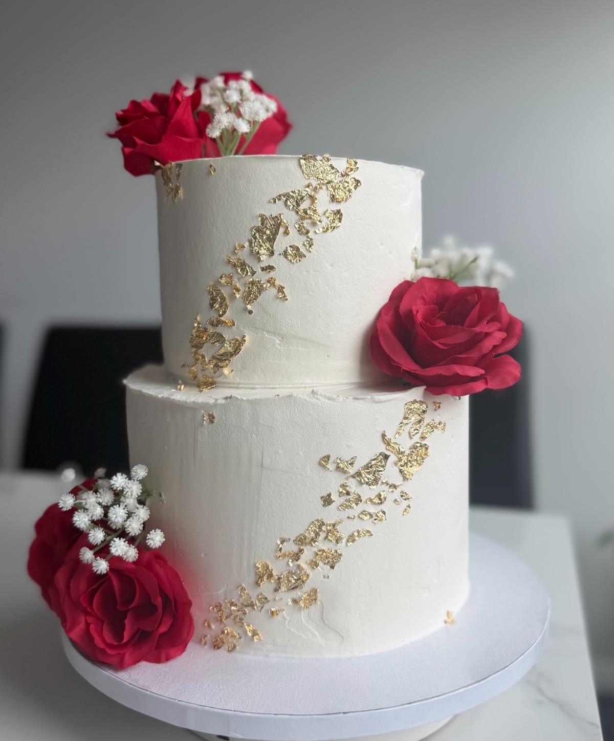 Two-tier white buttercream cake with a diagonal seam of gold leaf, topped and flanked with deep red roses and sprigs of baby's breath.