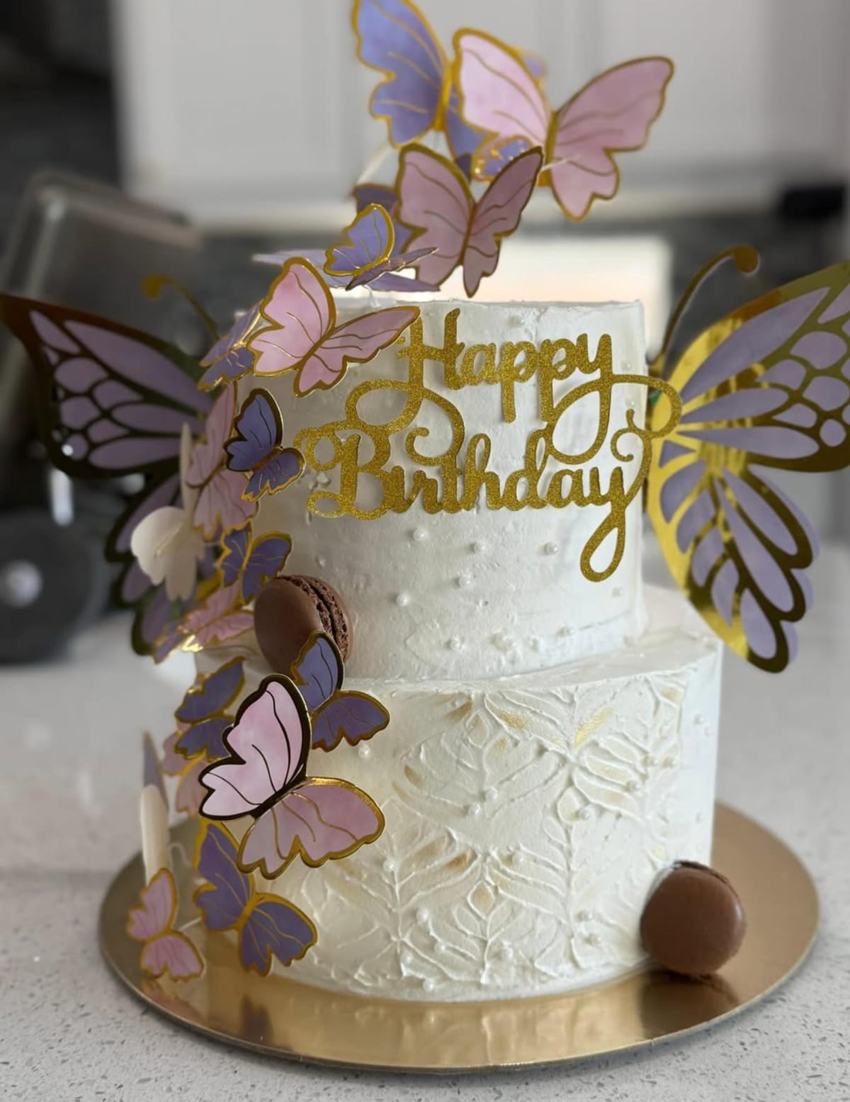 Two-tier white cake with pink and lavender butterflies climbing one side, large gold butterfly wings behind, a gold Happy Birthday topper and two macarons on the lower tier.