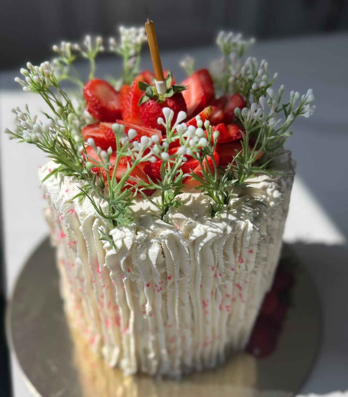 Tall cake with vertical piped buttercream ridges flecked with pink, crowned with sliced strawberries and sprigs of small white blossom, lit by direct sunlight.