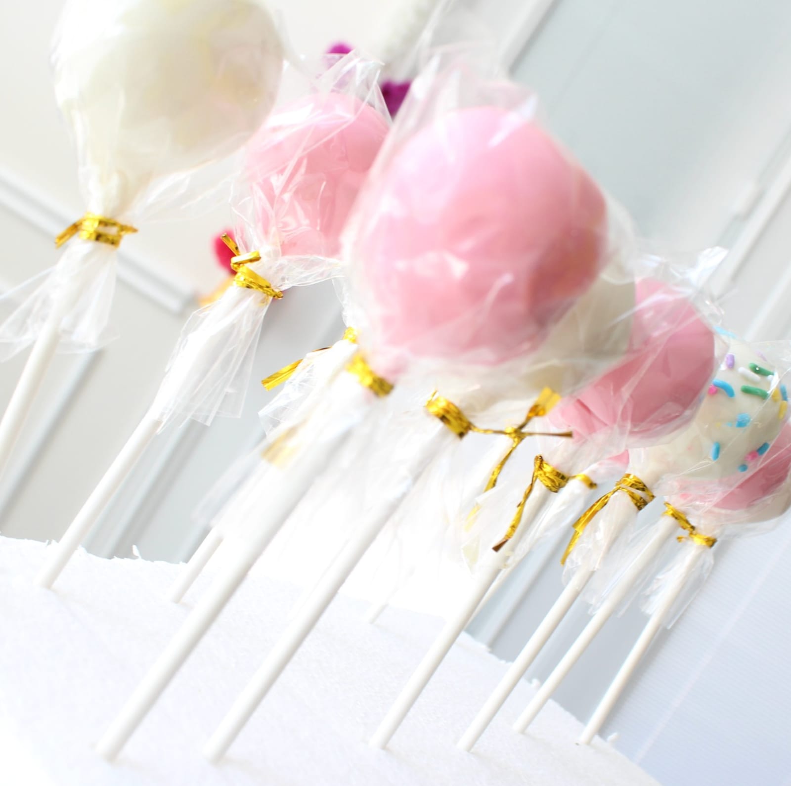 Cake pops in pink and white, each sealed in a clear cellophane bag tied with gold ribbon, standing upright on sticks in a white block.