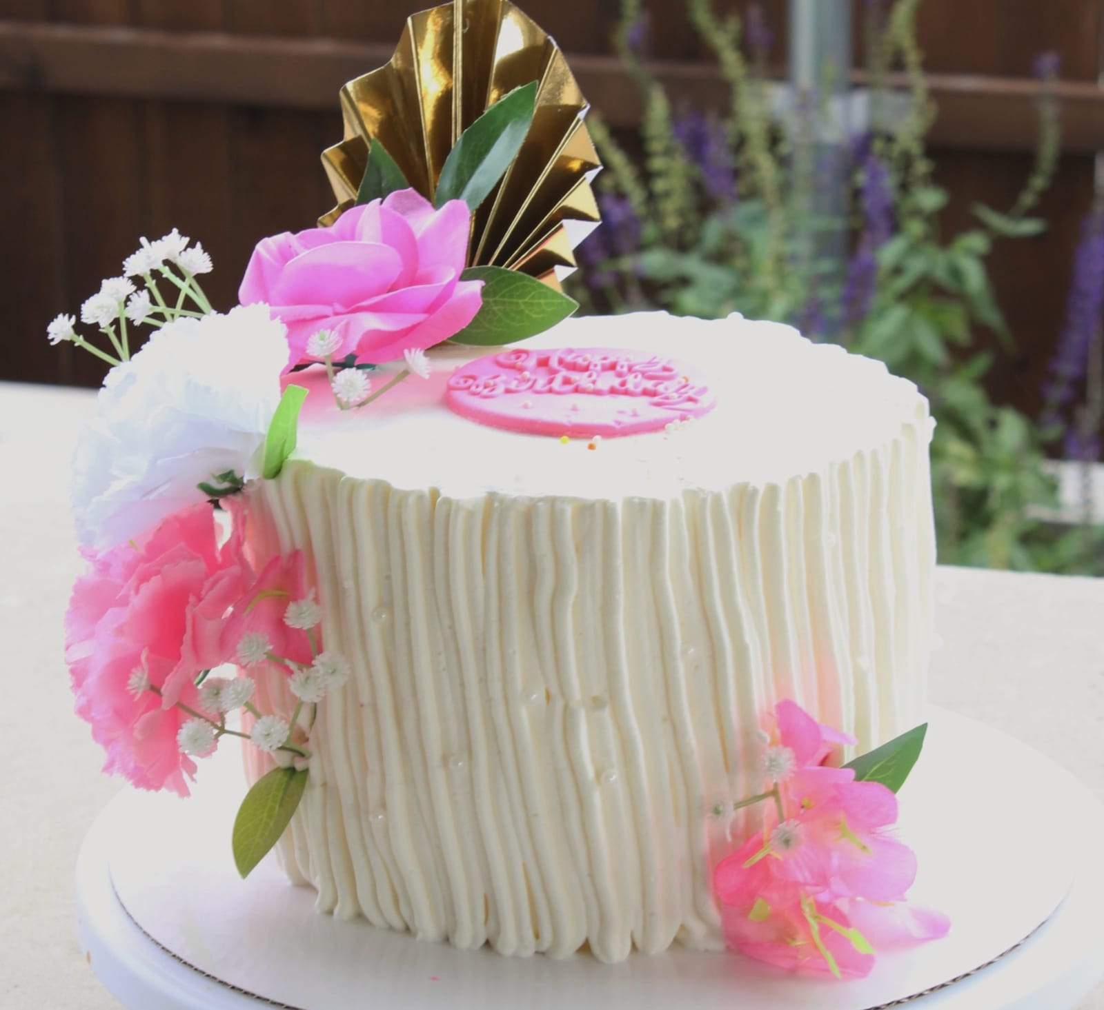 Single-tier cake with vertically combed ivory buttercream, pink and white flowers up one side, a gold pleated fan topper and a small pink plaque, photographed outdoors.